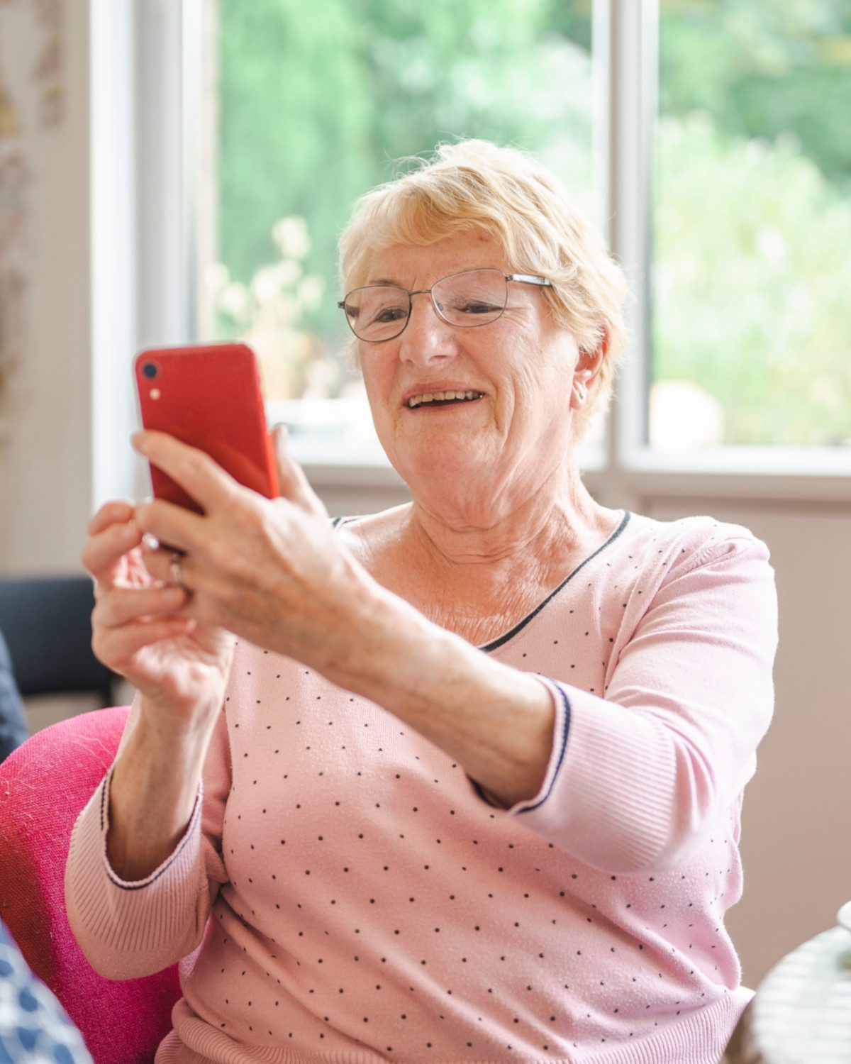 An older woman in glasses smiling warmly as she holds up her phone by a sunlit window.