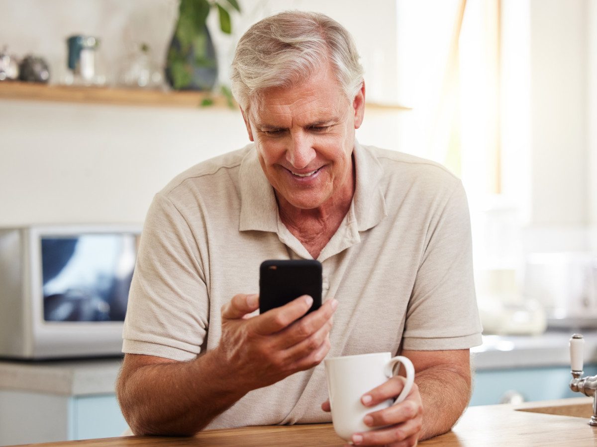 An older man smiling as he reads his phone in a bright kitchen, a mug of tea in his hand.