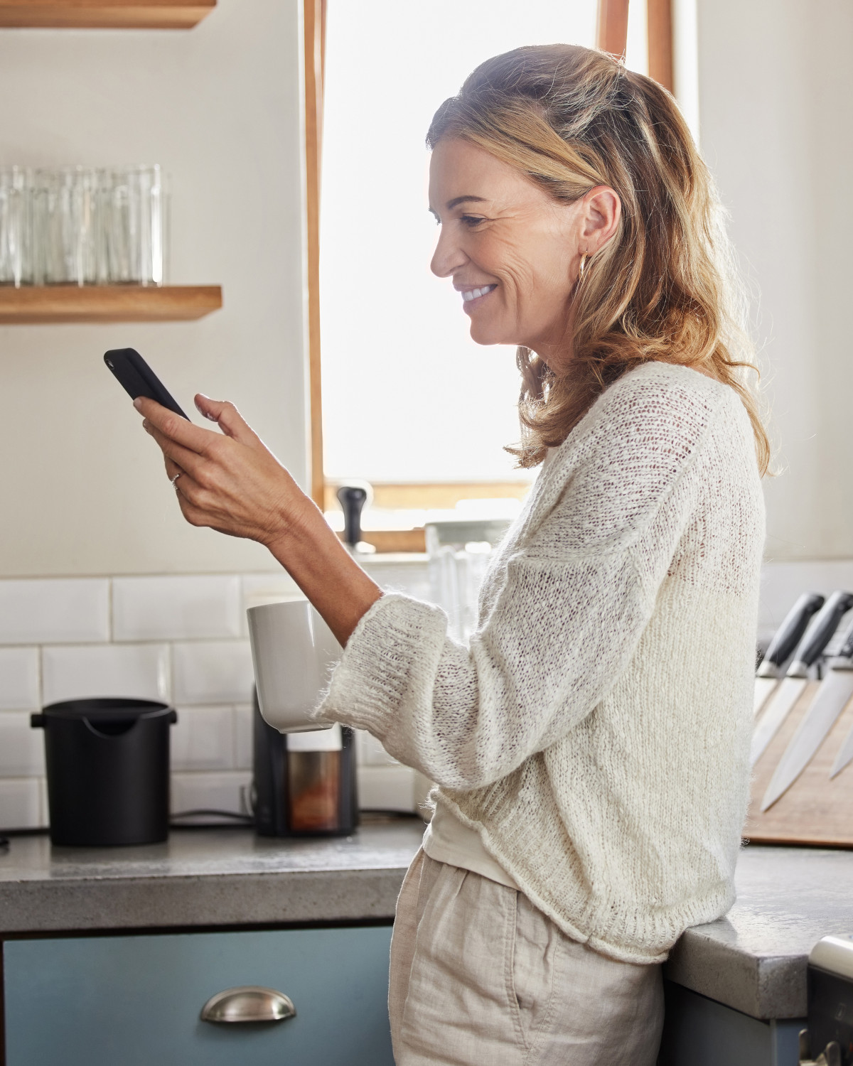 An older woman in a bright kitchen, smiling gently as she reads something on her phone.